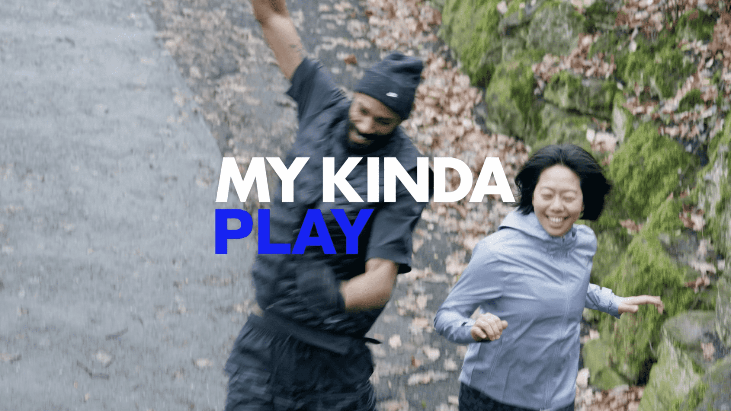 Two people jogging on a path surrounded by moss-covered rocks and autumn leaves. One wears a black top; the other a light blue jacket. Text overlay: "MY KINDA PLAY."